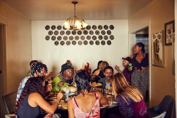 Guests at the party sit around two tables eating in front of an art piece that looks like the keys of a manual typewriter. One woman stands in the room’s doorway.