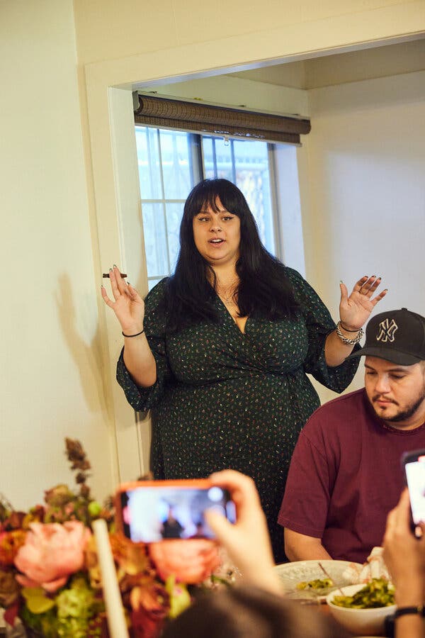A woman with a joint in her hand stands behind a man while talking to people at a table. A flower arrangement with cannabis buds is visible and people are holding up phones to take pictures.